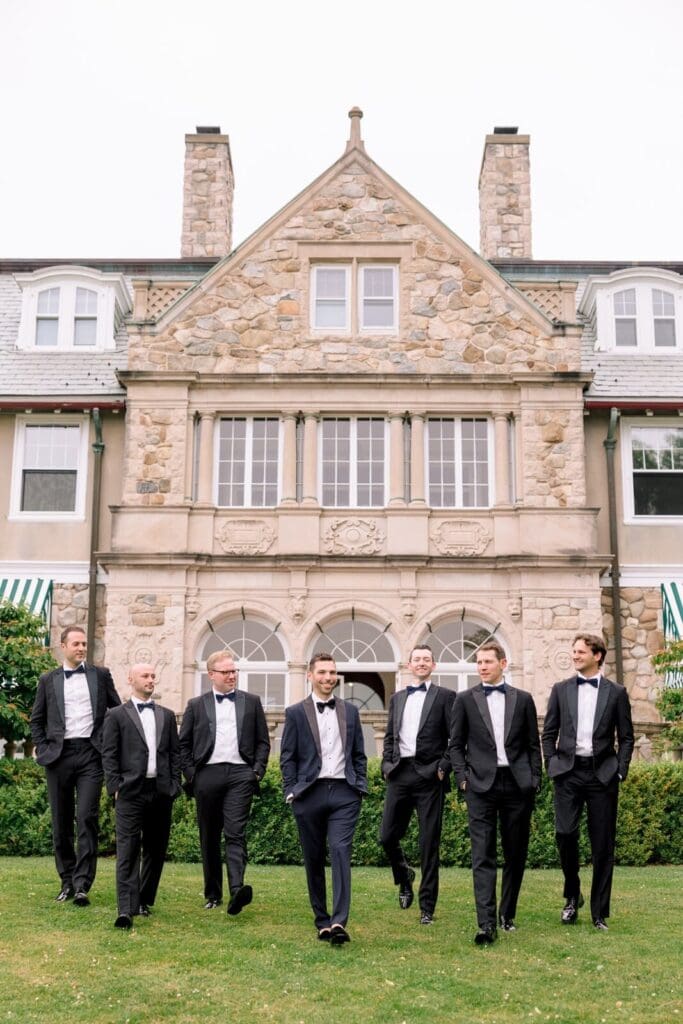 A groom and his wedding party walking in their tuxedos in front of a stone building wedding venue